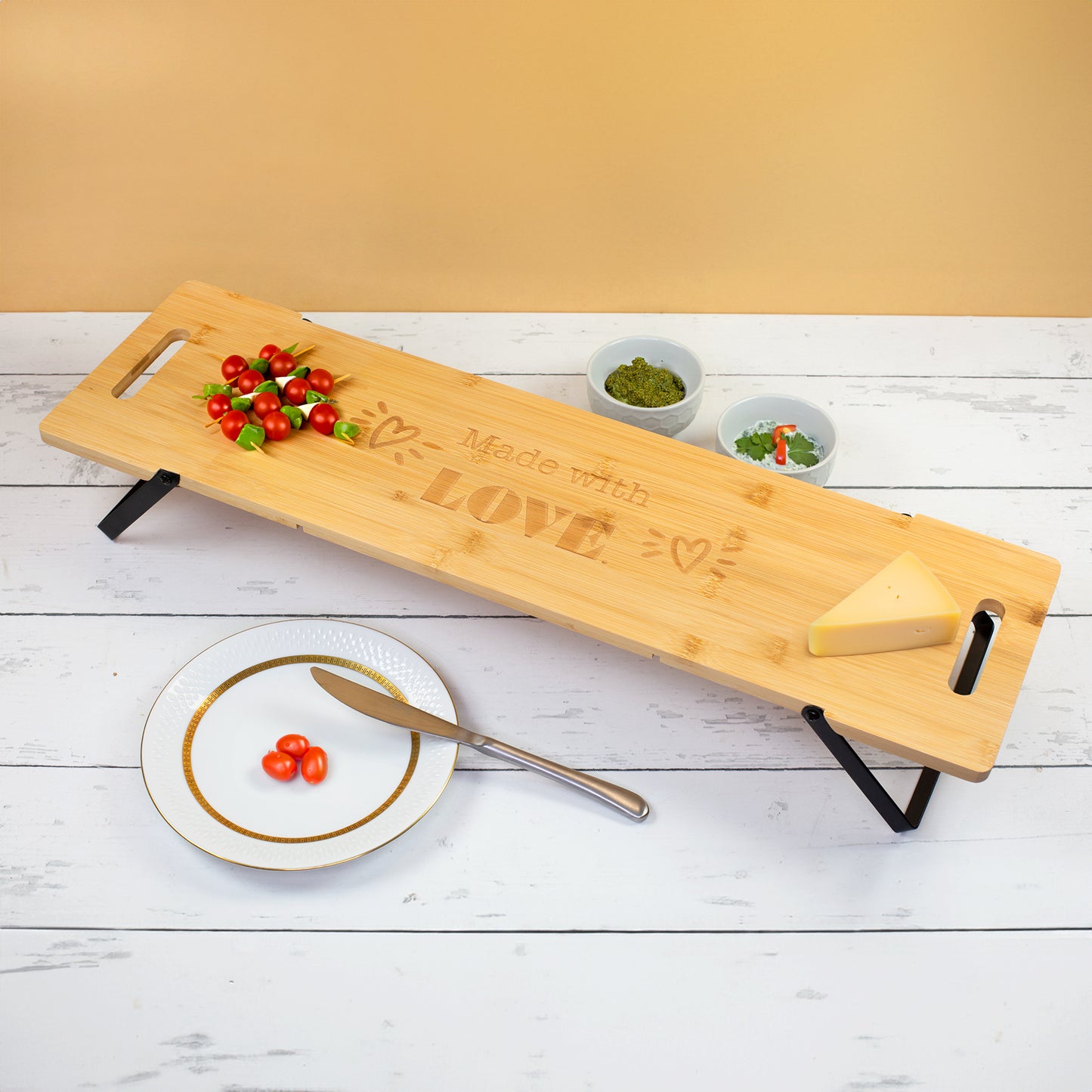 Großes Servierbrett aus zertifiziertem Bambus mit klappbaren Metallfüßen und Made with Love Gravur, angerichtet mit Tomaten, Käse, Dips und Snacks