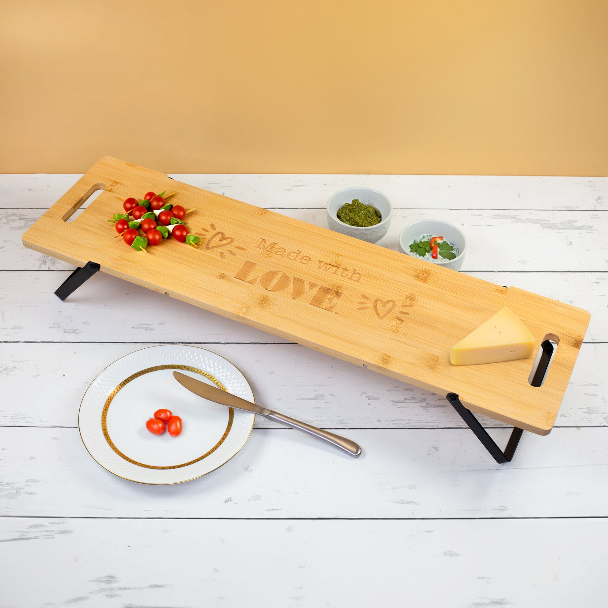 Großes Servierbrett aus zertifiziertem Bambus mit klappbaren Metallfüßen und Made with Love Gravur, angerichtet mit Tomaten, Käse, Dips und Snacks