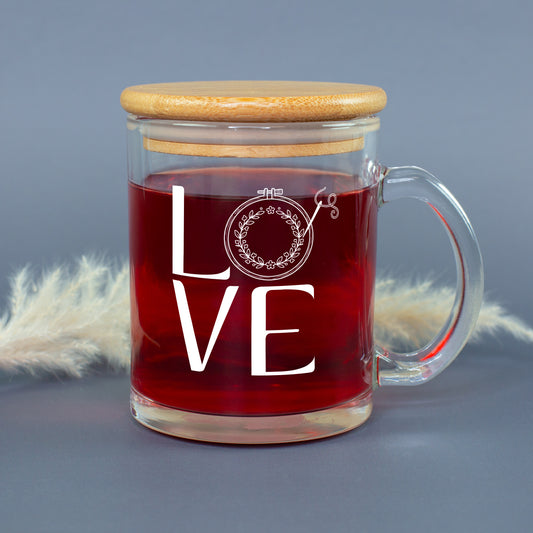 Glastasse mit rotem Tee, LOVE Stickmotiv und aufgesetztem Bambusdeckel vor grauem Hintergrund