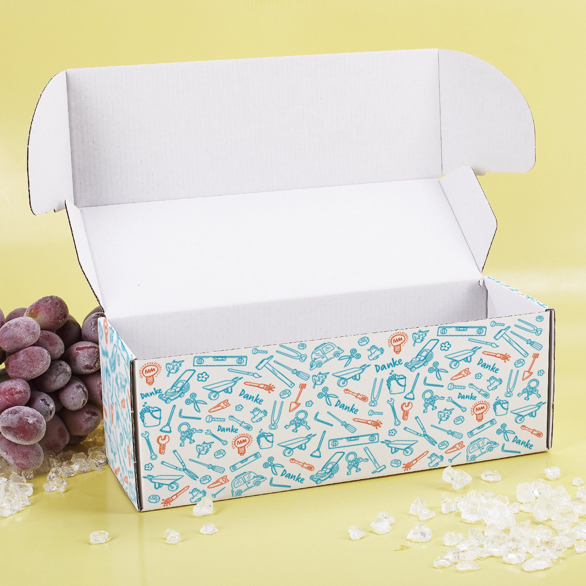 Geöffnete Geschenkbox mit farbigem Werkzeug-Muster und Deko auf gelbem Hintergrund mit WeintraubenDeko.
