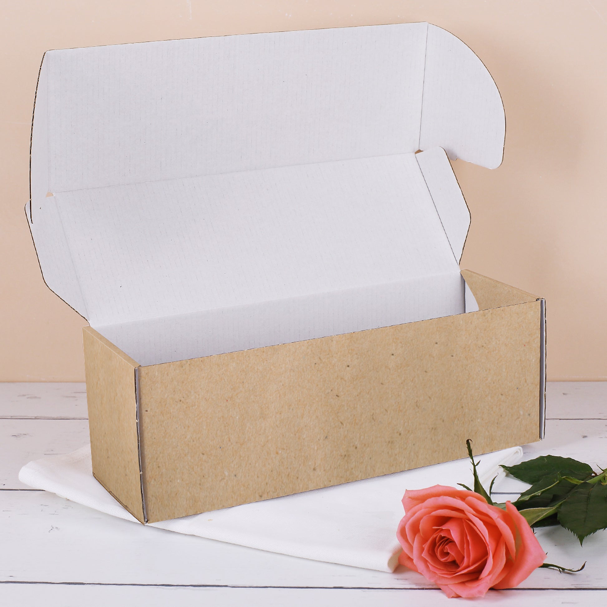 Geöffnete längliche Geschenkbox im Kraftpapier-Stil – innen weiß, daneben eine rosafarbene Rose