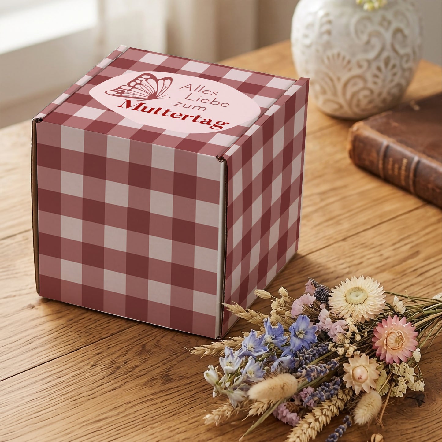 Muttertags Geschenkbox mit rosa Karomuster und Schmetterlingsmotiv auf Holztisch neben Trockenblumen