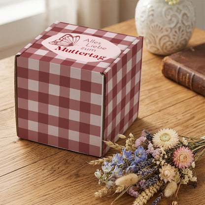 Muttertags Geschenkbox mit rosa Karomuster und Schmetterlingsmotiv auf Holztisch neben Trockenblumen