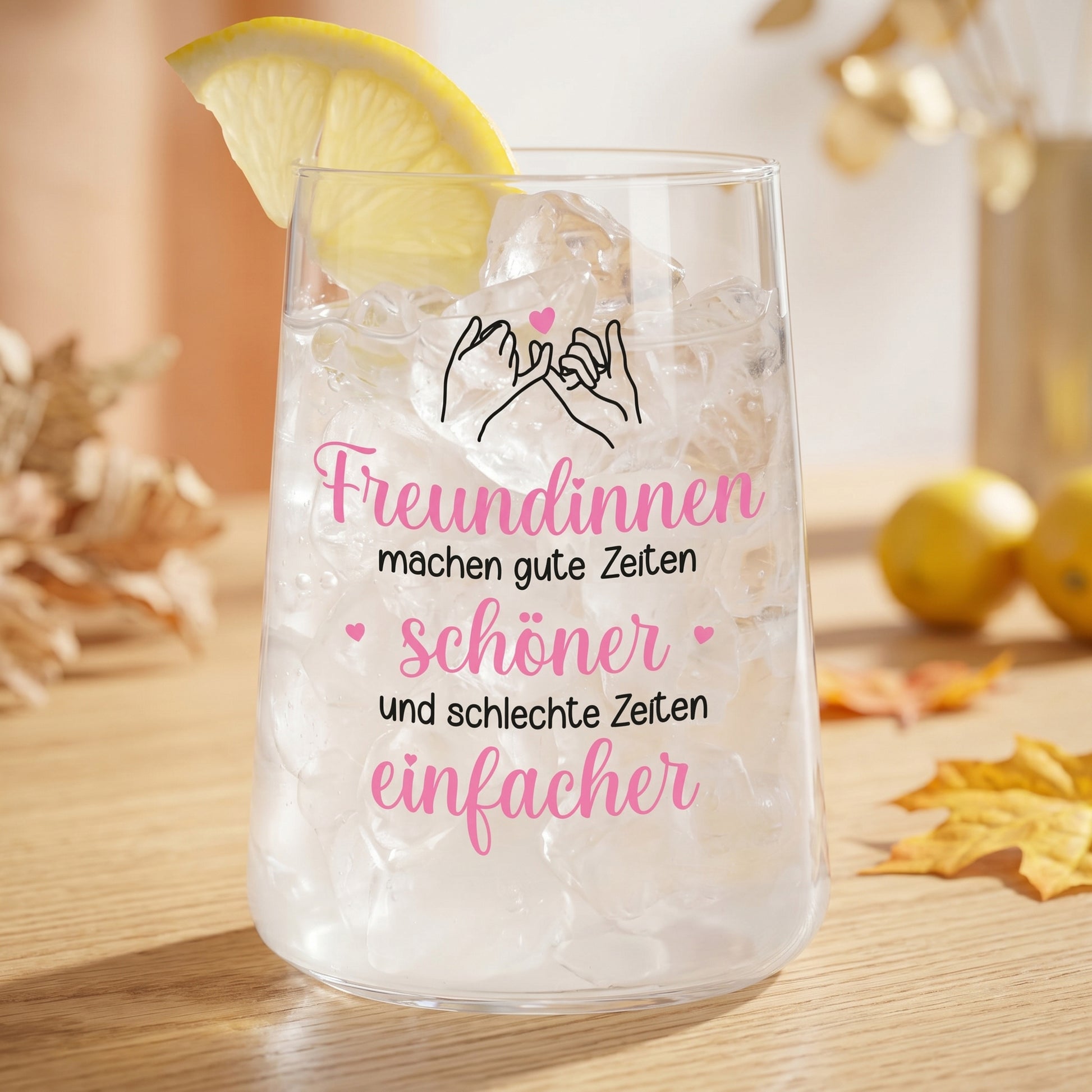 Trinkglas mit Eiswürfeln, Zitronenscheibe und Freundinnen-Motiv auf hellem Tisch