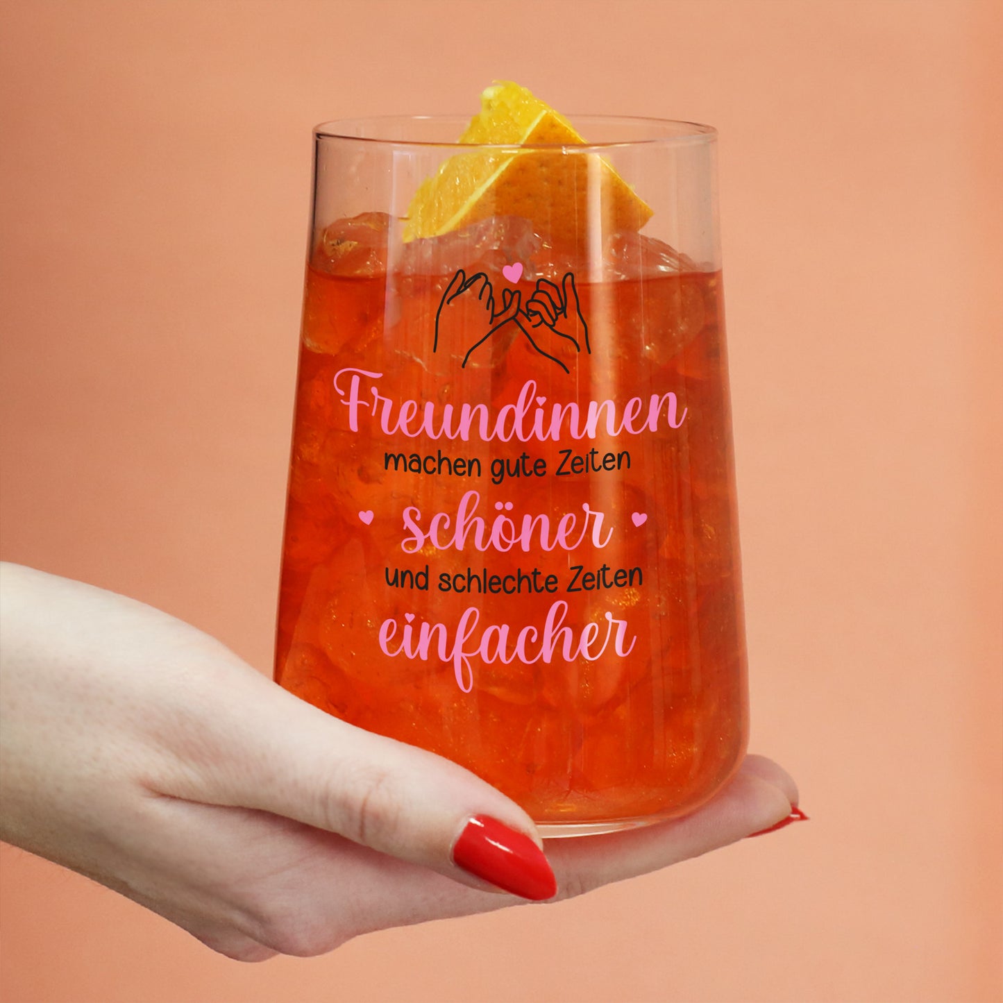 Trinkglas mit rotem Getränk, Eiswürfeln, Orangenscheibe und Freundinnen-Motiv in der Hand gehalten