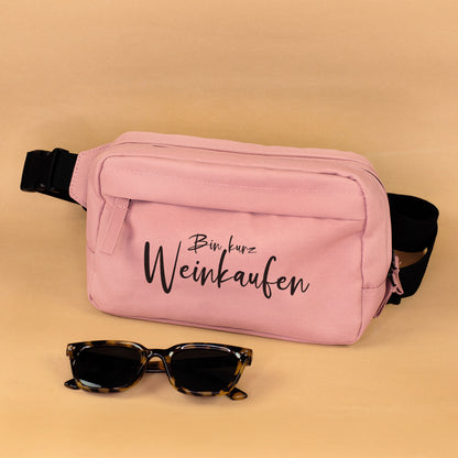 Rosa Bauchtasche mit Spruch Bin kurz Weinkaufen und schwarzem Gurt mit Sonnenbrille als Deko