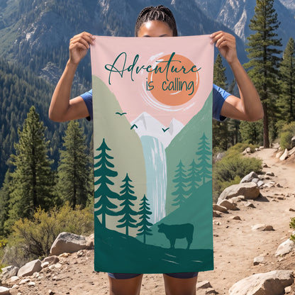 Person hält Sporthandtuch mit Abenteuer-Motiv vor Berglandschaft, Aufdruck mit Bergen, Wasserfall und Schriftzug Adventure is calling