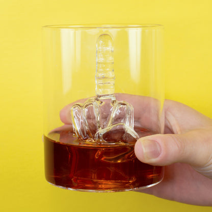 Mittelfinger Whiskyglas in der Hand mit dunklem Getränk und gelbem Hintergrund