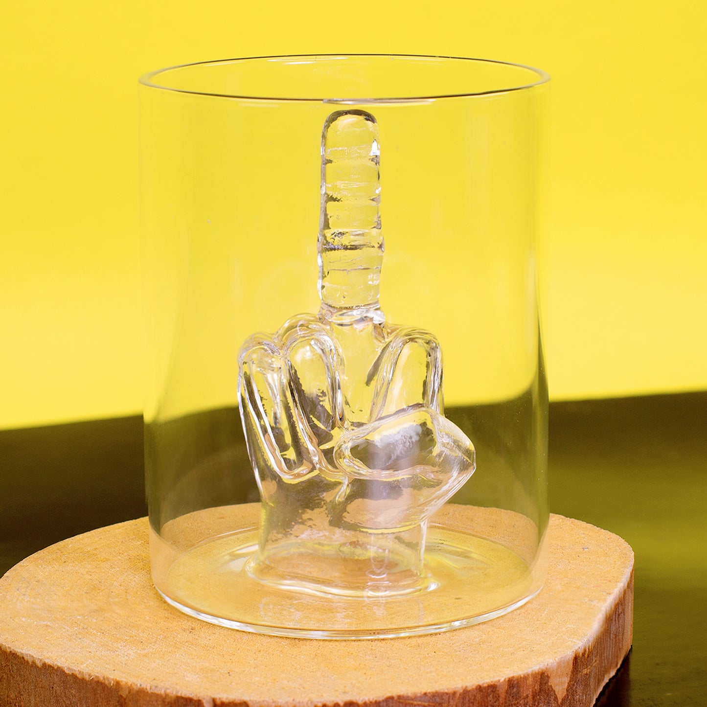 Mittelfinger Trinkglas aus transparentem Glas auf Holzscheibe vor gelbem Hintergrund