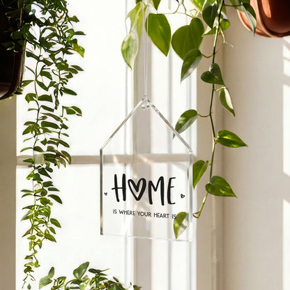 Transparenter HOME Acrylanhänger in Hausform hängt am Fenster zwischen grünen Pflanzen