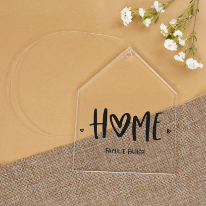Transparenter Acrylanhänger in Hausform mit HOME-Aufdruck und personalisiertem Text Familie Faber auf Stoffunterlage mit weißen Blumen