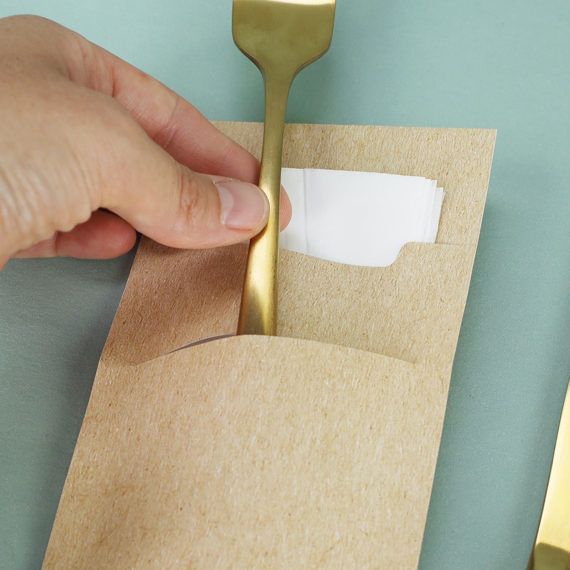 Detailaufnahme einer Bestecktasche aus naturbraunem Papier mit goldener Gabel und Serviette