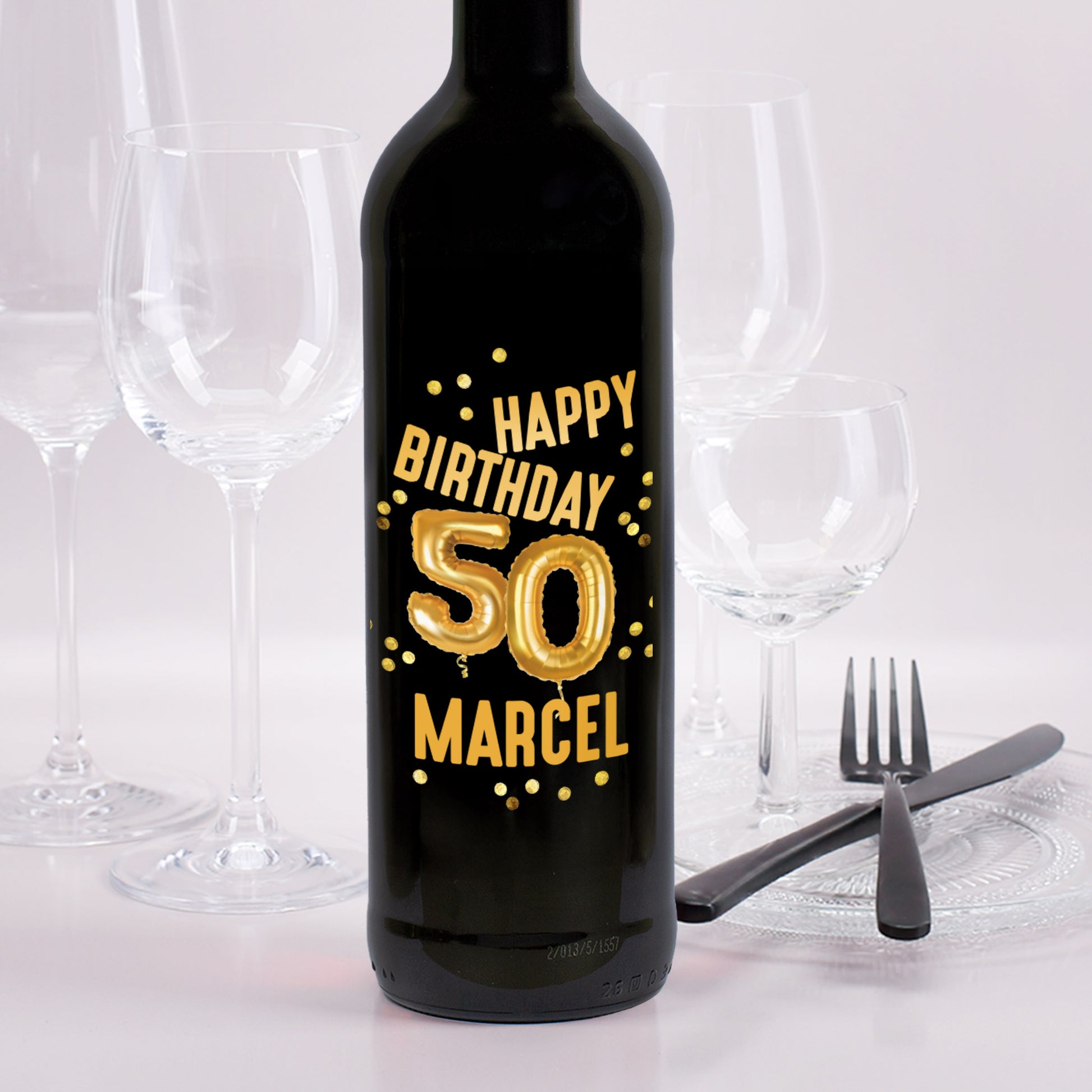 Schwarze Weinflasche mit goldener Ballonzahl „50“ und dem Text „Happy Birthday Marcel“ auf dem Etikett, daneben leere Weingläser und Besteck