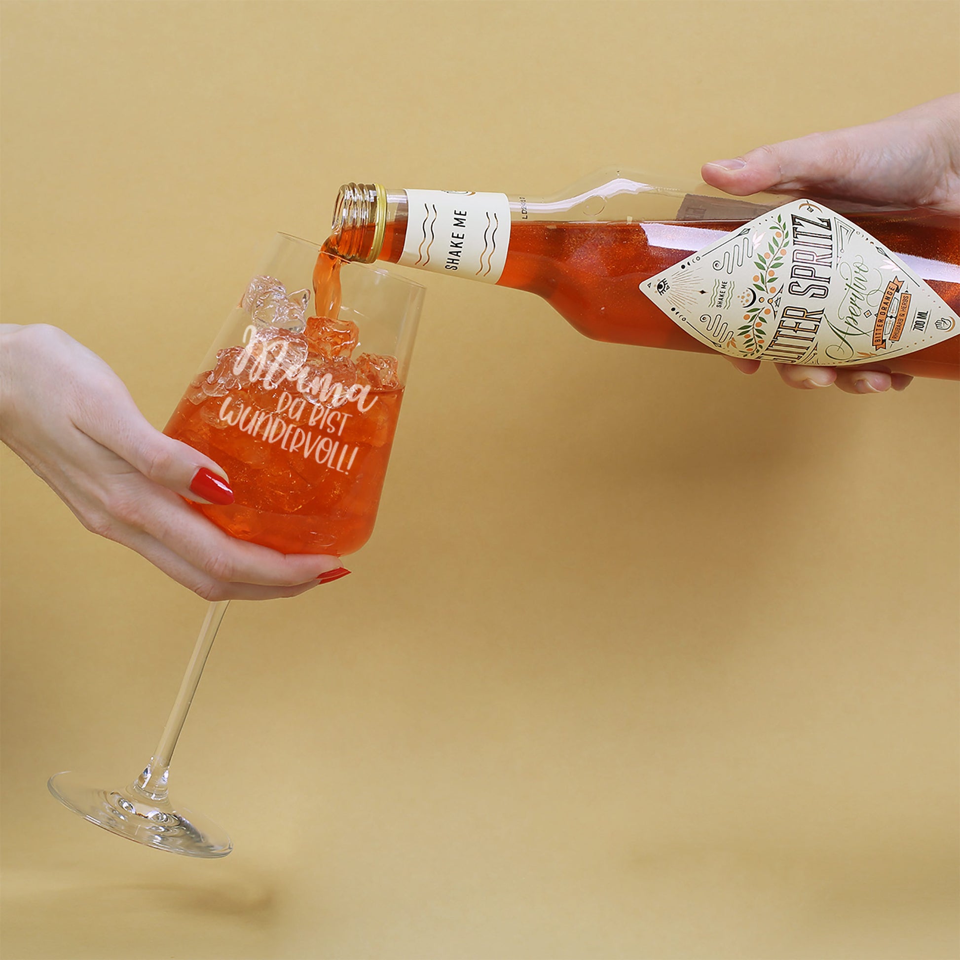 Hand gießt Glitter Spritz in ein Cocktailglas mit Gravur „Mama, du bist wundervoll!“ auf hellem Untergrund