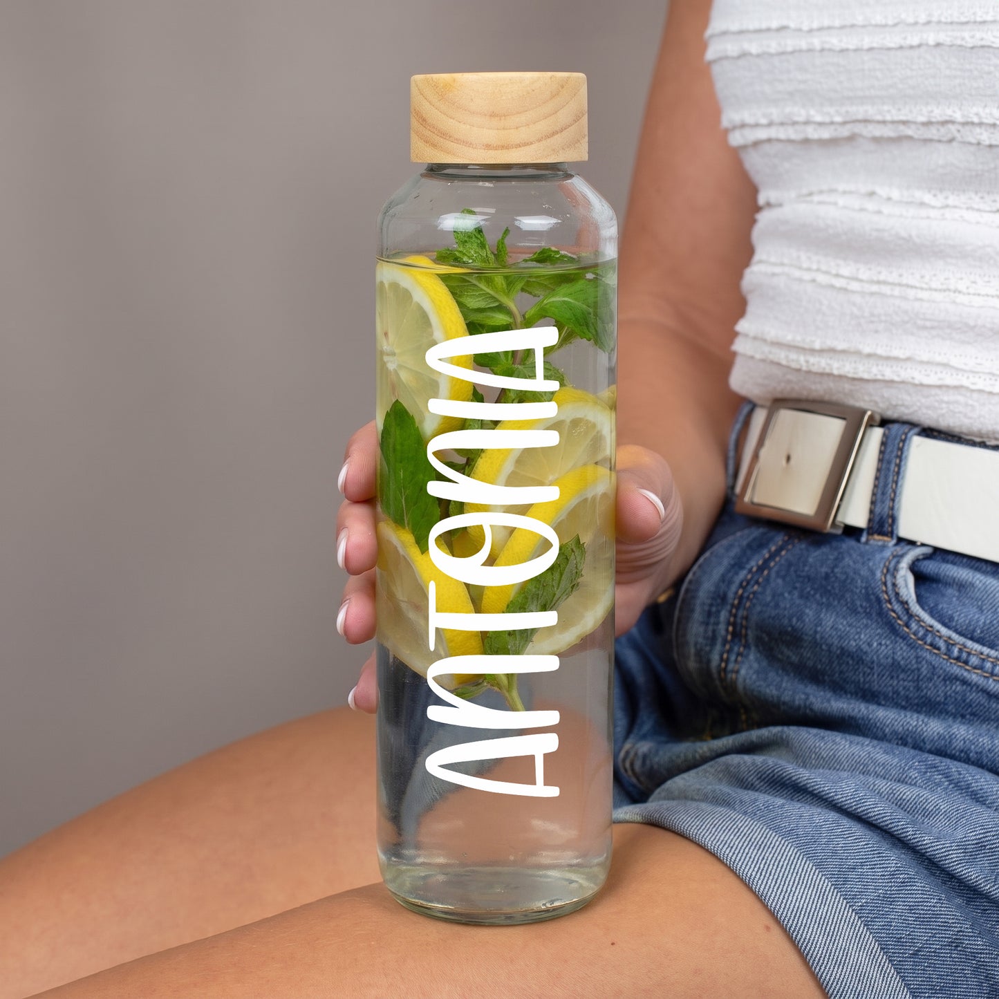 Handgehaltene Trinkflasche aus Glas mit weißem Namensaufdruck Antonia, gefüllt mit Wasser, Zitronenscheiben und Minze, mit Holzdeckel