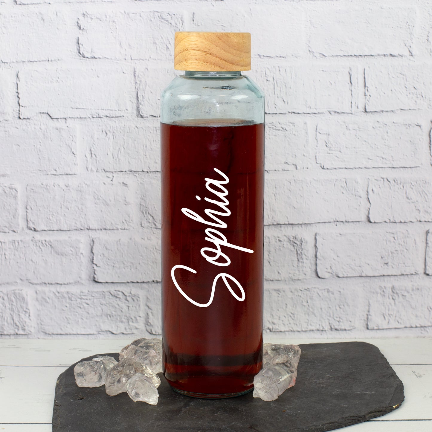 Personalisierte Trinkflasche aus recyceltem Glas mit weißem Namen Sophia, Holzdeckel und dunklem Getränk auf Schieferplatte