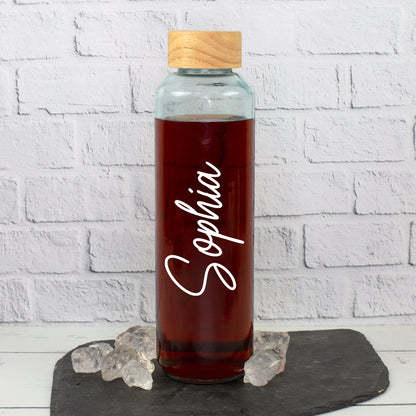 Personalisierte Trinkflasche aus recyceltem Glas mit weißem Namen Sophia, Holzdeckel und dunklem Getränk auf Schieferplatte