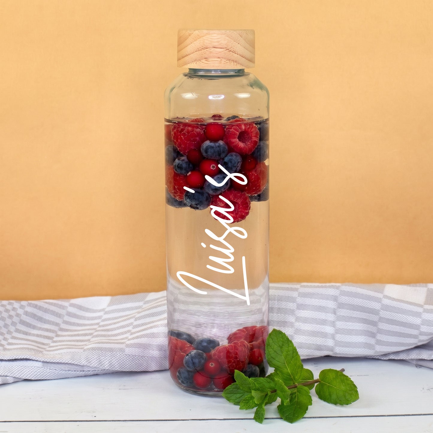 Trinkflasche aus Glas mit weißem Namen Luisa’s, gefüllt mit Wasser, Beeren und Minze als Infused Water