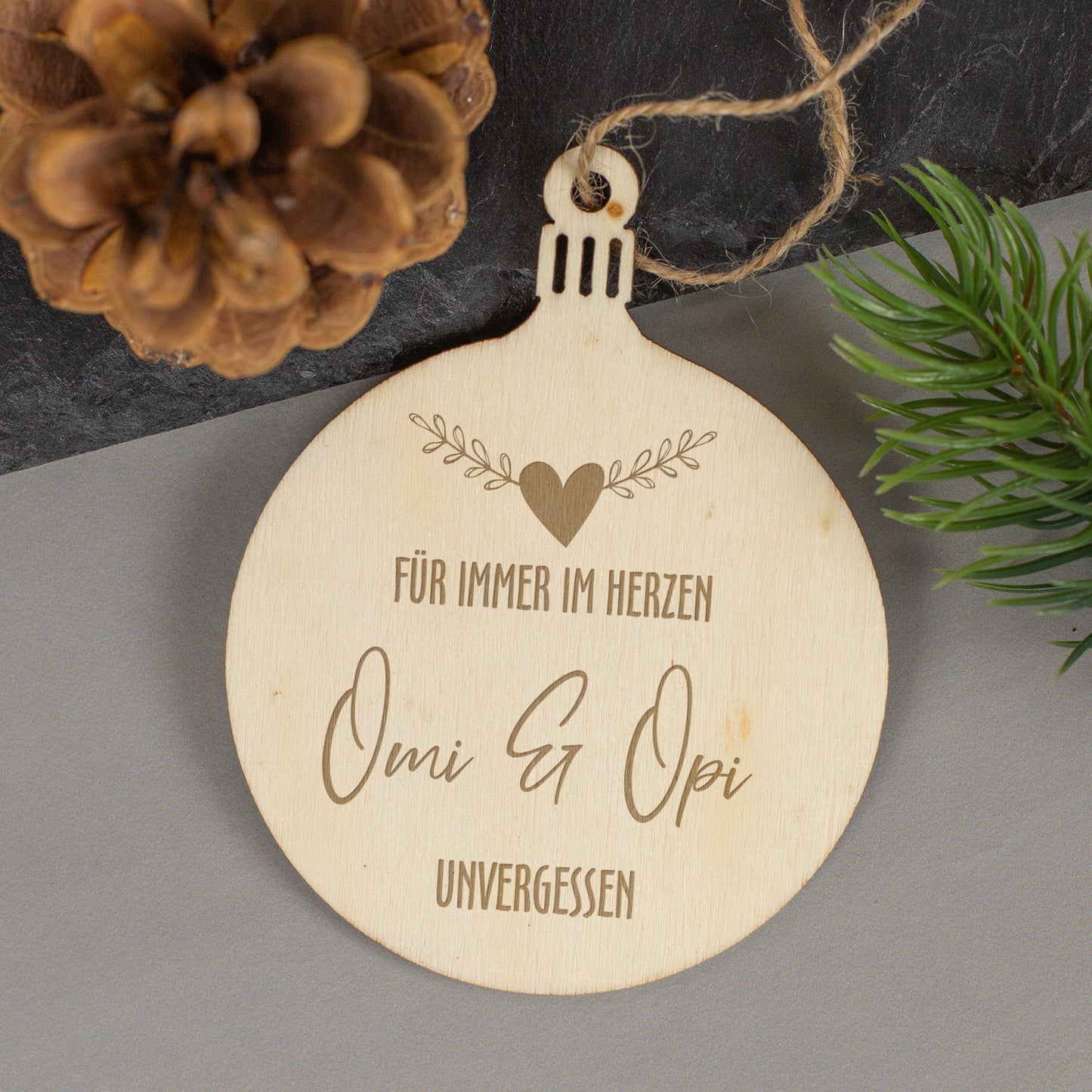 Gedenkanhänger aus Holz mit Gravur „Für immer im Herzen“, personalisiert mit Inschrift Omi & Opi, umgeben von Tannenzapfen und Zweigen.