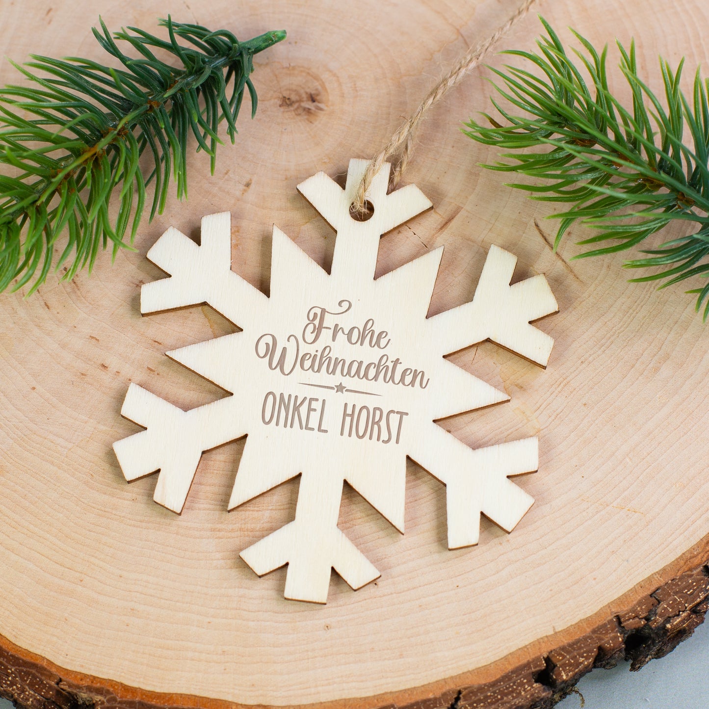 Personalisierter Holzanhänger Schneeflocke mit Gravur „Frohe Weihnachten Onkel Horst“, liegend auf Holzscheibe mit Tannenzweigen.