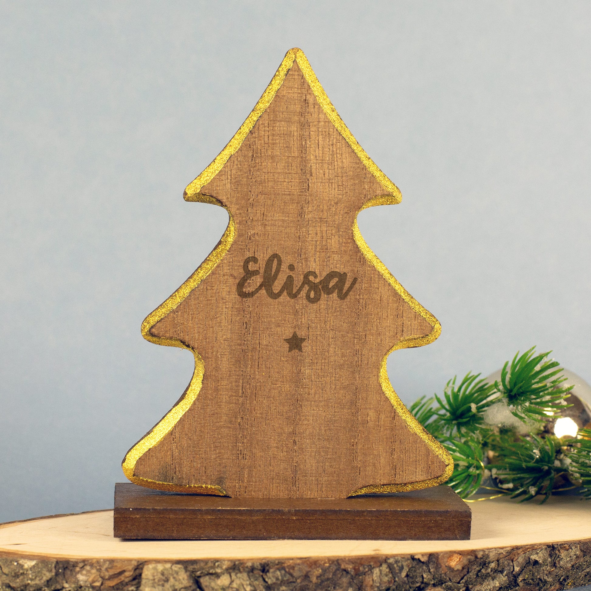 Holz-Tannenbaum mit goldenem Rand und graviertem Namen „Elisa“, darunter ein kleiner eingravierter Stern, auf Holzsockel stehend, dekoriert mit Tannenzweig und Lichterkette.