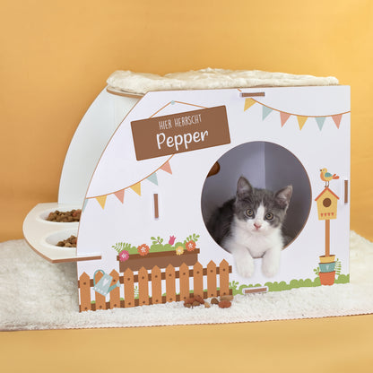 Weißes Katzenhaus mit Gartenmotiv, personalisiertem Namen Pepper, runder Türöffnung, zwei Näpfen und Katze im Innenbereich