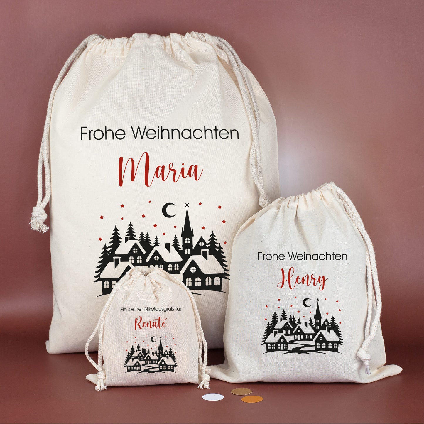 Drei Geschenksäcke in verschiedenen Größen mit Winterdorf-Motiv, roten Sternen und individuellem Wunschtext – festlich arrangiert auf bordeauxfarbenem Hintergrund.
