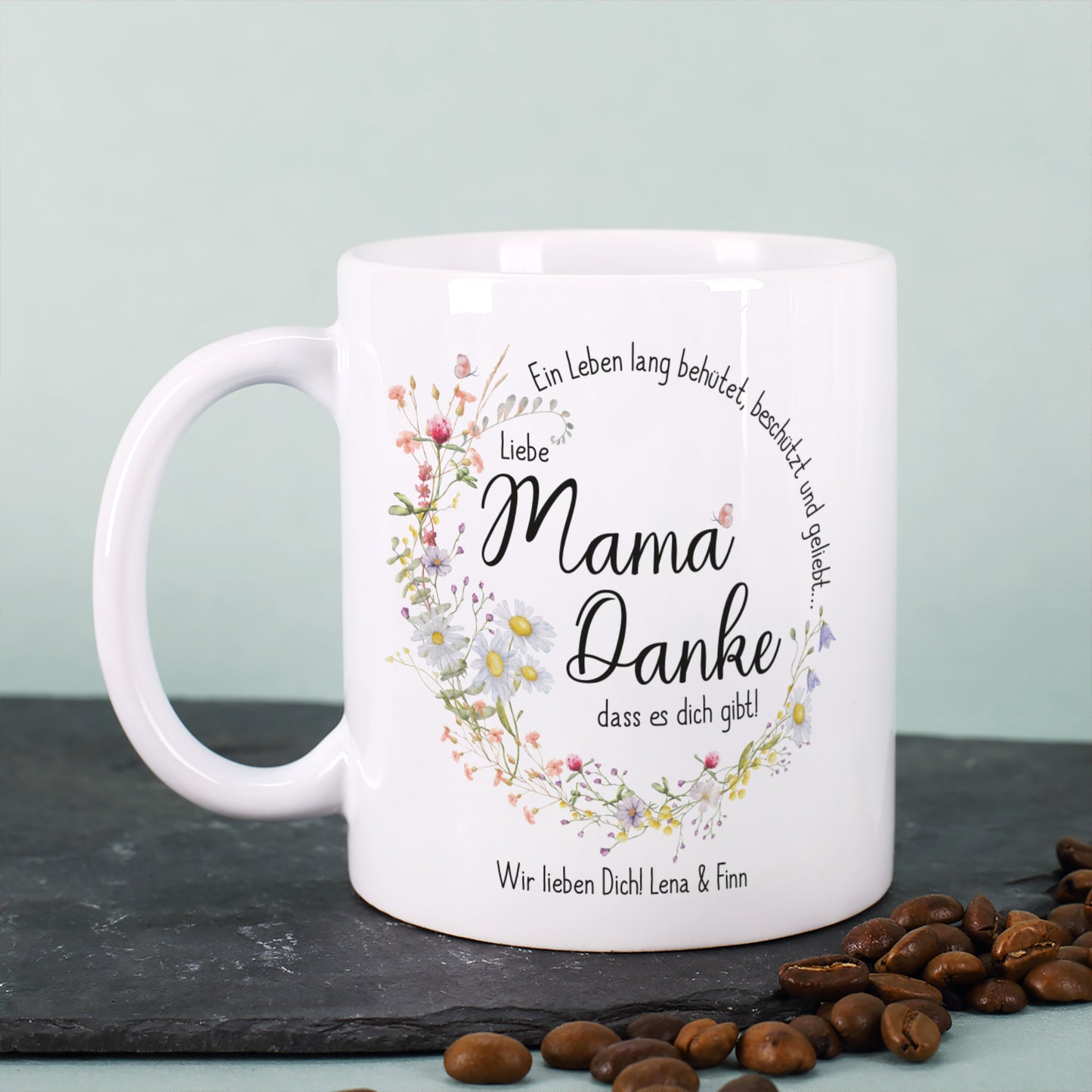 Weiße Keramiktasse mit seidenglänzendem Finish, bedruckt mit einem kreisförmigen Wiesenblumen-Kranz und dem Spruch „Ein Leben lang behütet, beschützt und geliebt…“. In der Mitte steht „Liebe Mama, danke, dass es dich gibt!“, ergänzt durch einen personalisierten Wunschtext. Die Tasse steht auf einer dunklen Steinplatte, umgeben von Kaffeebohnen, mit einem sanft grünen Hintergrund. Ein liebevolles Geschenk für Mütter zum Muttertag, Geburtstag oder als Dankeschön.