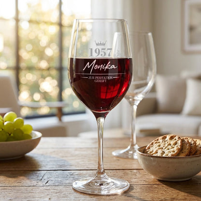 Leonardo Weinglas mit Gravur „1957 Monika – Zur Perfektion gereift“, mit Rotwein gefüllt auf Holztisch neben Trauben und Gebäck