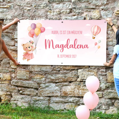 Rosa Geburtsbanner mit Teddy Motiv, Name Magdalena und Wunschtext „Hurra, es ist ein Mädchen“, von zwei Personen an Steinwand gehalten