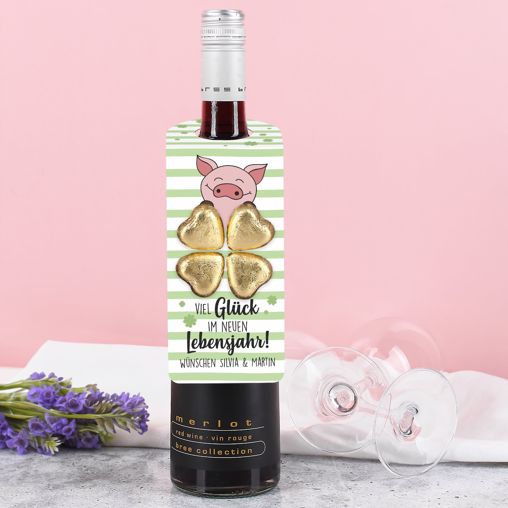 Flaschenanhänger mit grün-weißem Streifendesign, Kleeblättern, Glücksschwein und vier goldenen Lindt Schokoladenherzen, an einer Flasche befestigt.