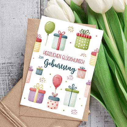 Personalisierte Glückwunschkarte zum Geburtstag mit Geschenken, Ballons und Umschlag neben weißen Tulpen