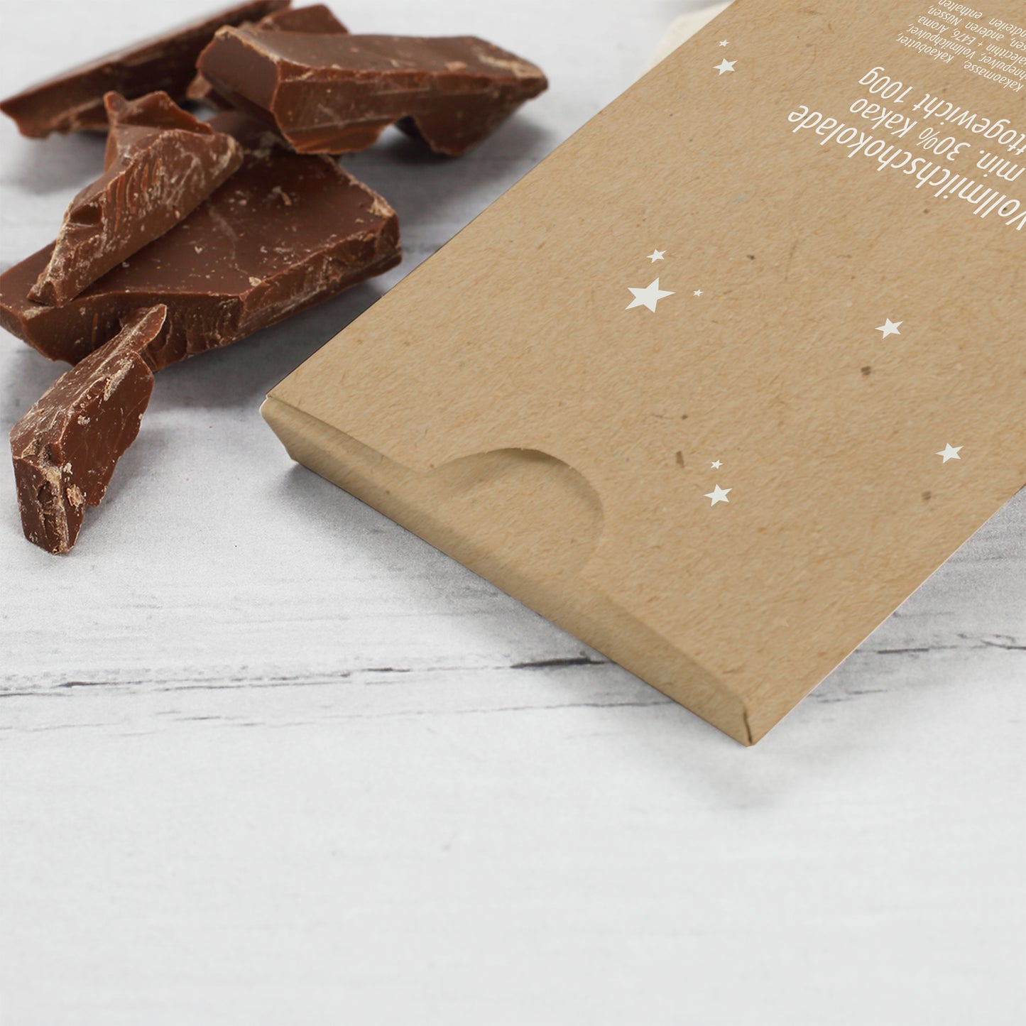 Rückseite der personalisierten Weihnachts-Schokolade mit feinem Sternenmuster auf braunem Kraftpapier. Im Bild liegen Stücke zarter Vollmilchschokolade neben der Verpackung, auf der die Nährwertangaben und Produktinformationen sichtbar sind. Detailaufnahme der hochwertigen Verarbeitung und natürlichen Materialstruktur der Verpackung.