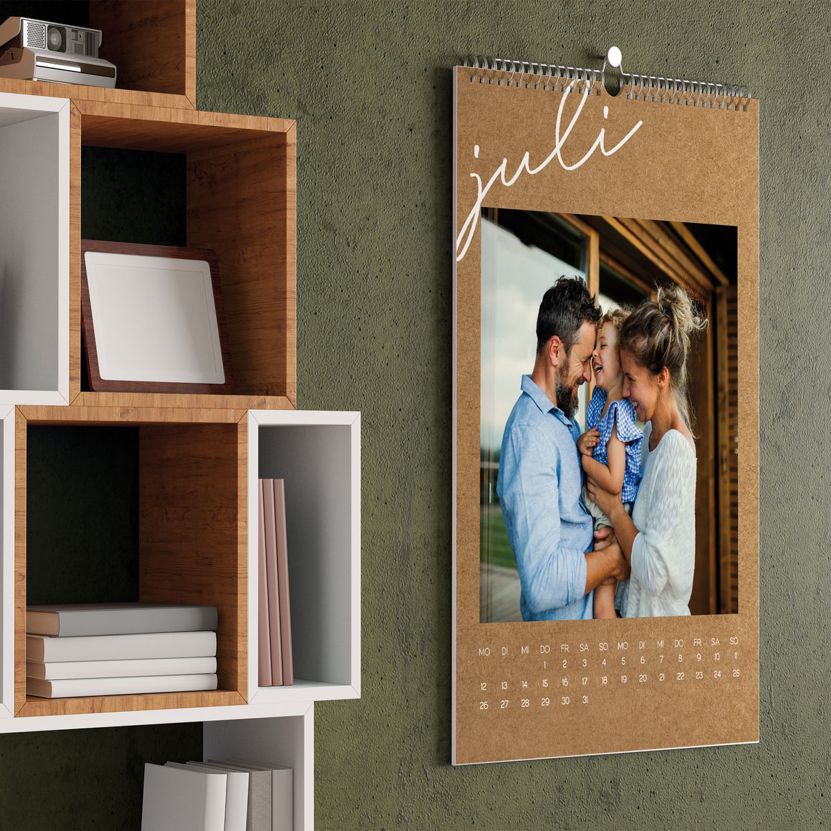 Foto-Wandkalender 2027 in Kraftpapier-Optik für den Monat Juli, mit Familienfoto und feiner Spiralbindung, aufgehängt an einer grünen Wand.