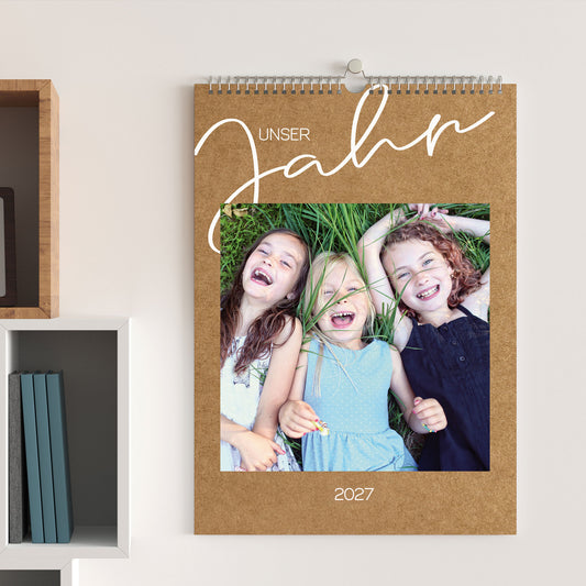 Foto-Wandkalender 2027 in Kraftpapier-Optik, aufgehängt an einer hellen Wand, mit Foto von drei lachenden Kindern und dem Titel „Unser Jahr“.
