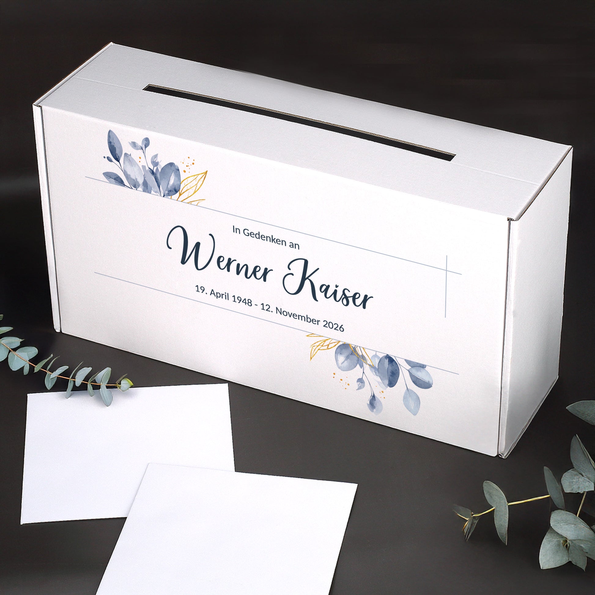Personalisierte weiße Briefbox für Trauerfeiern mit Blattmotiv, Name und Datum, stabiler Karton, seitlicher Schlitz, dekoriert mit Eukalyptuszweigen.