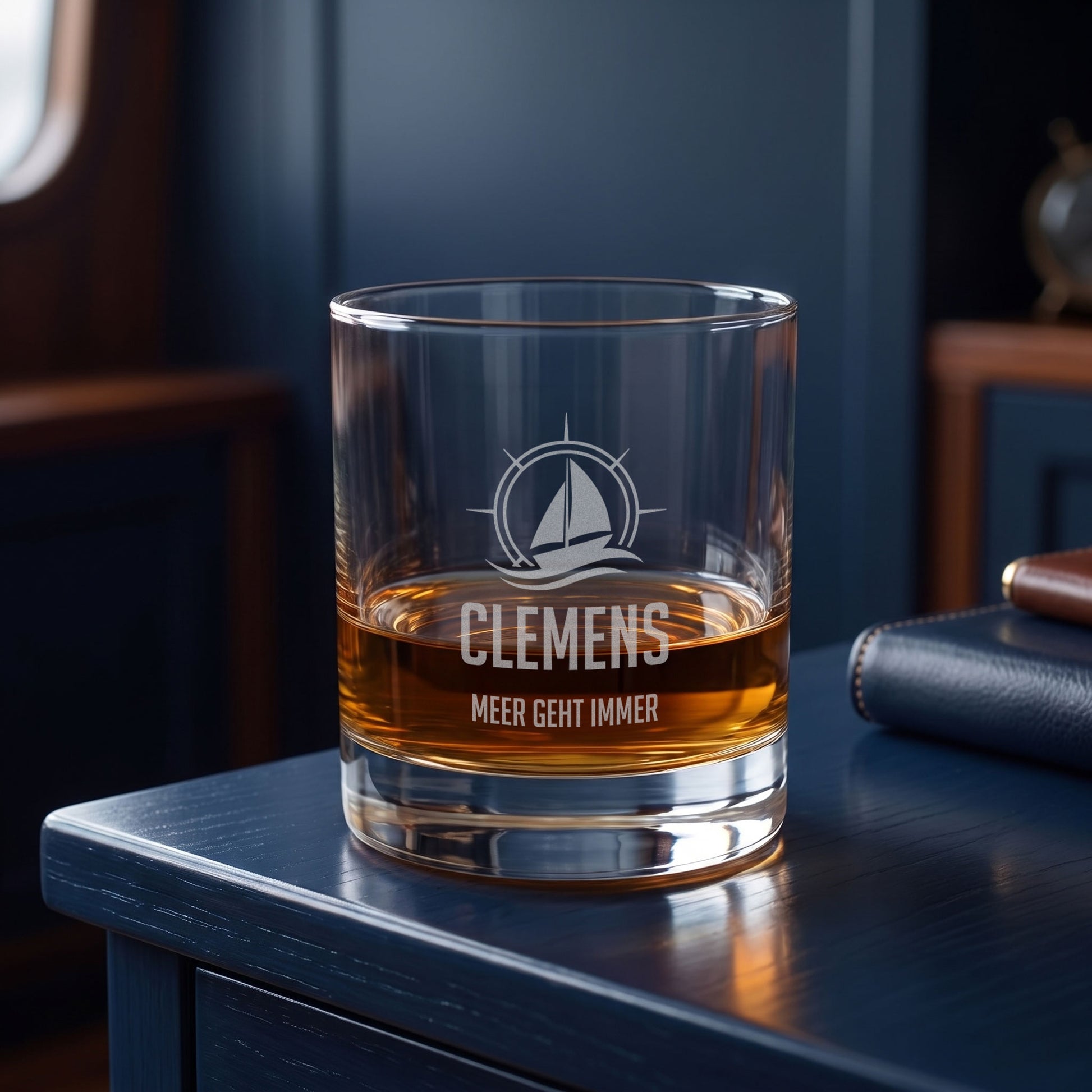 Whiskeyglas mit Segelboot-Gravur, Name Clemens und Wunschtext Meer geht immer auf blauem Tisch