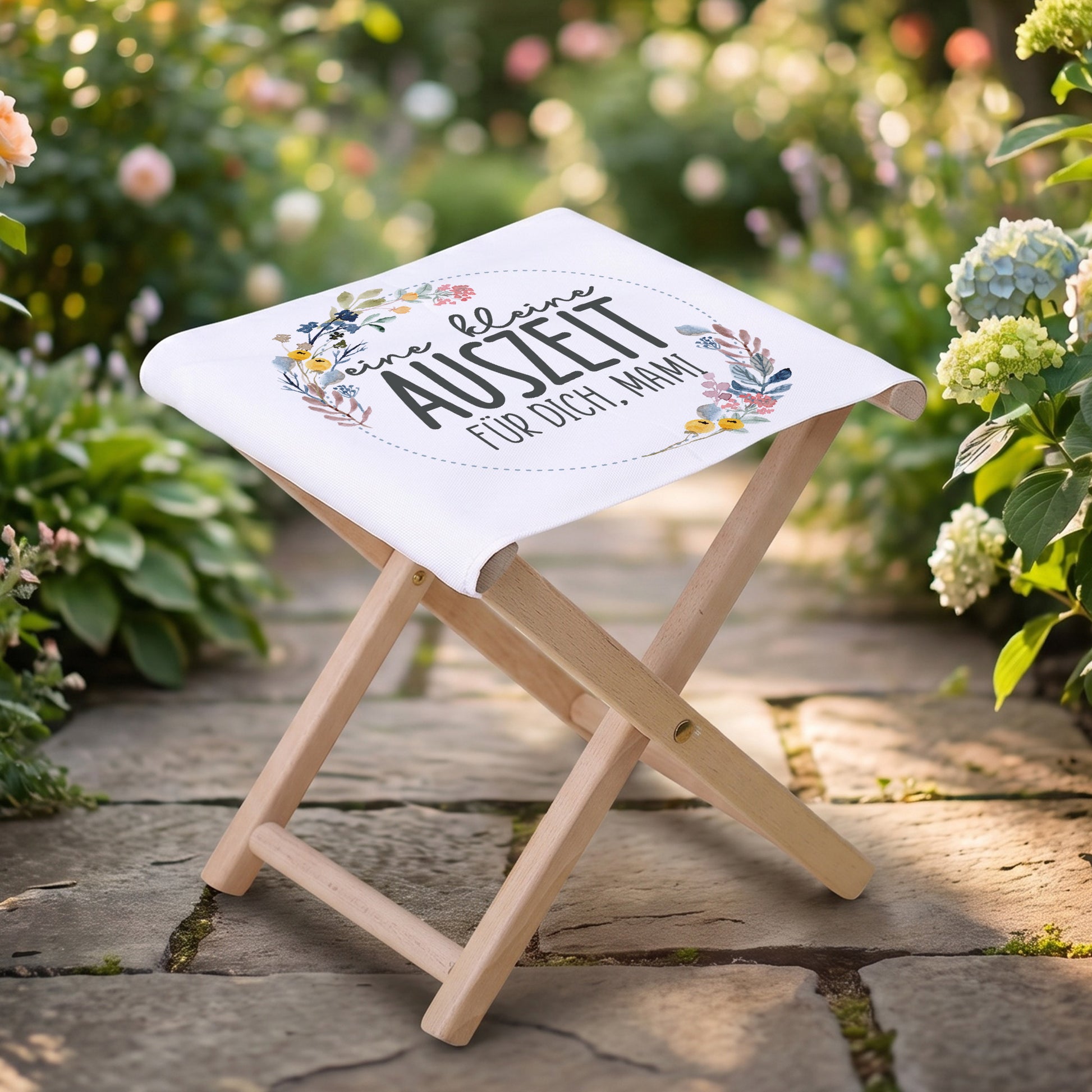 Klapphocker mit Aufdruck „eine kleine Auszeit für dich, Mama“ im Gartenweg platziert