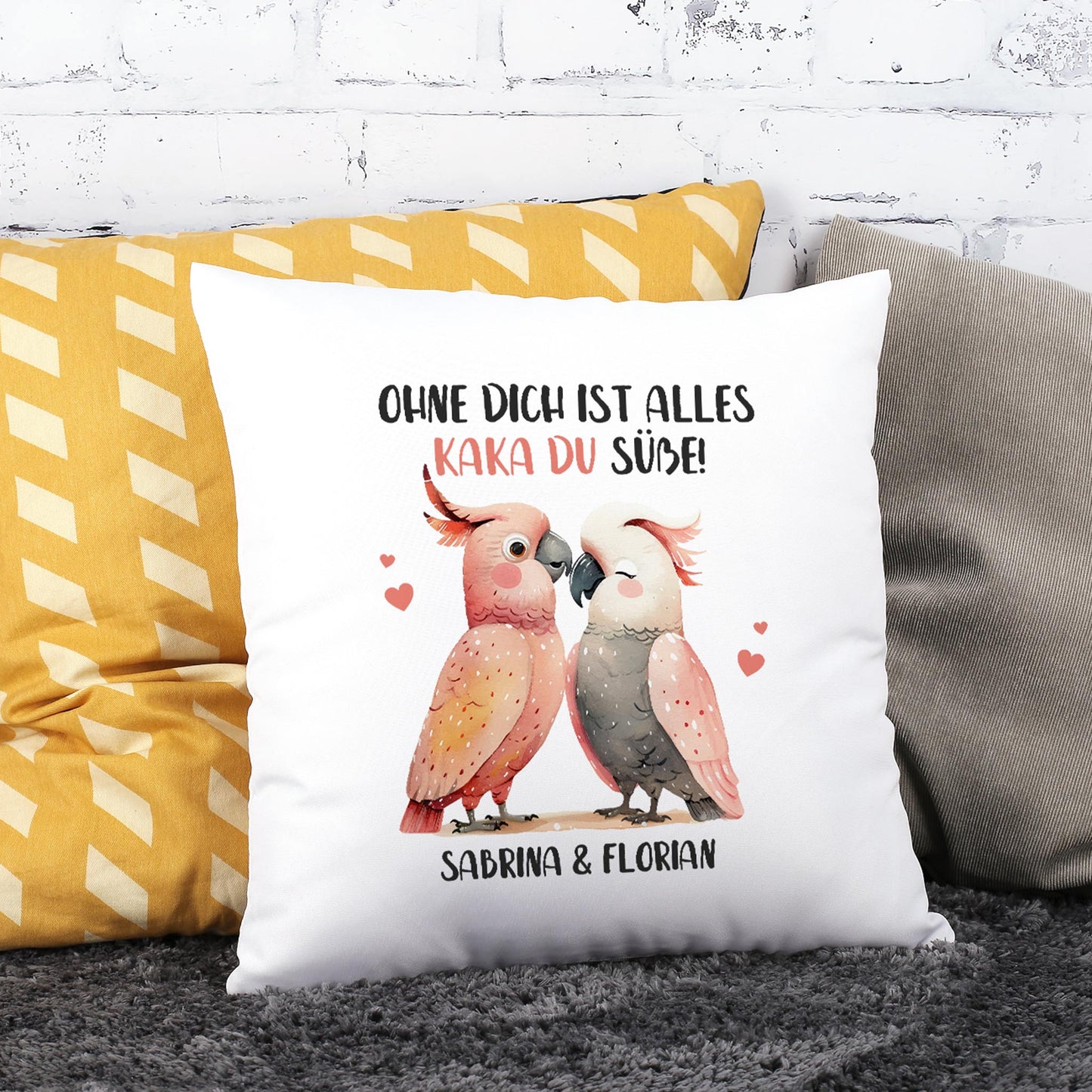 Personalisiertes Kakadu-Kissen mit romantischem Spruch und zwei Namen, dekoriert auf Sofa vor Backsteinwand