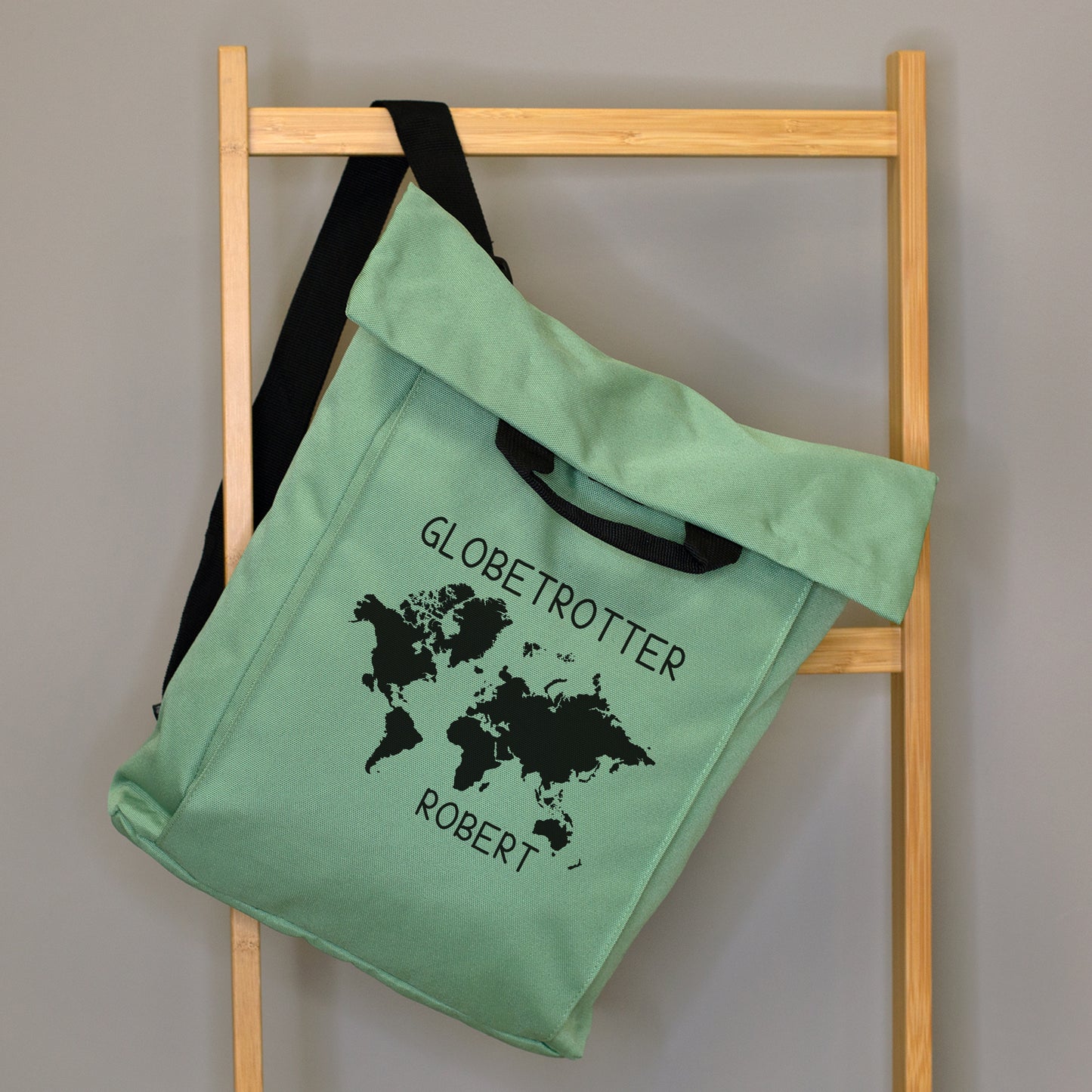 Mintgrüner 2-in-1 Rucksack mit Weltkarten-Motiv, Wunschtext „Globetrotter“ und Name „Robert“ an einer Holzleiter aufgehängt