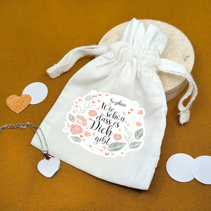 Bedruckter Geschenkbeutel mit Blumenmotiv und Name – Spruch „Wie schön, dass es dich gibt“ – auf Geschenkunterlage mit Herzanhänger