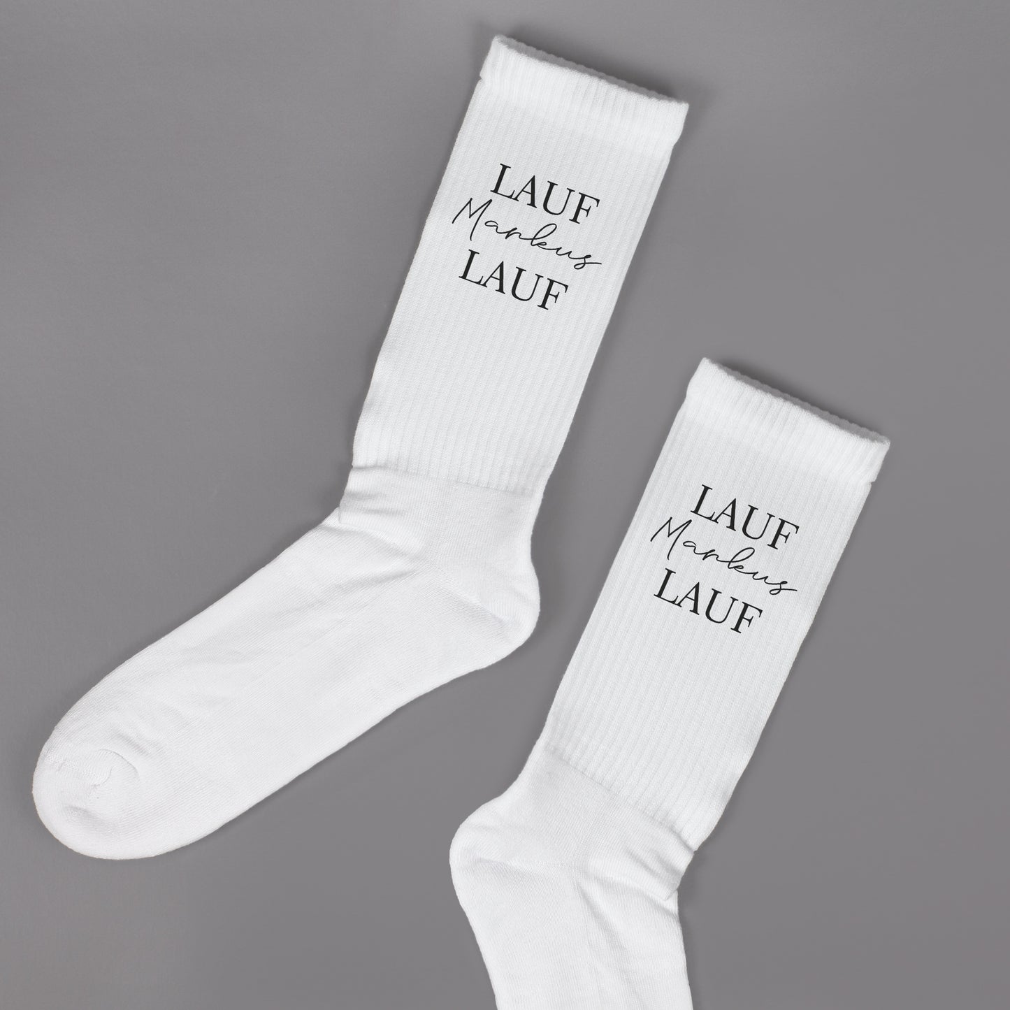 Paar weiße Sportsocken mit personalisiertem „Lauf Markus Lauf“-Aufdruck auf grauem Hintergrund