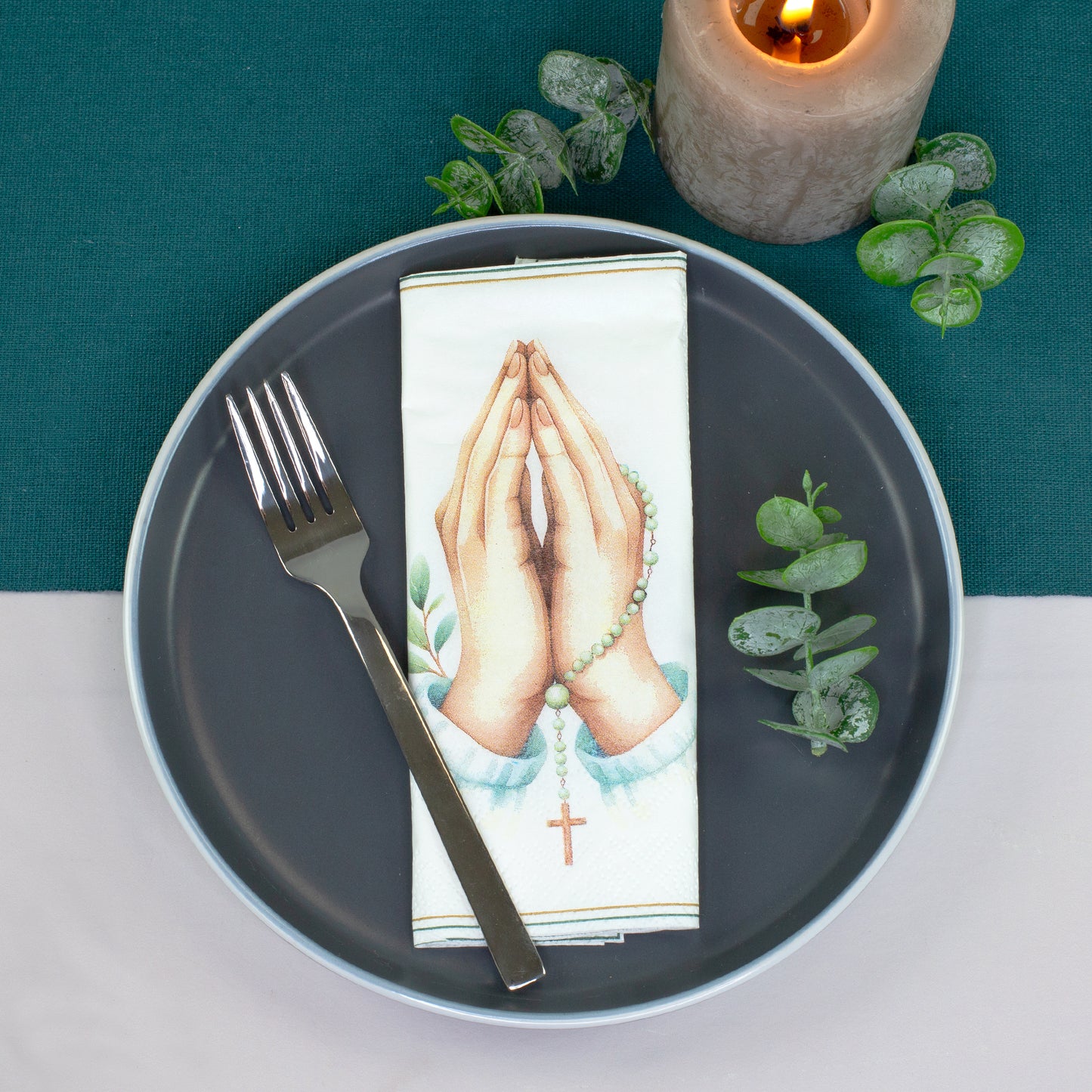 Gefaltete Serviette mit Gebetshände Motiv auf festlich gedecktem Teller mit Gabel, Kerze und Eukalyptus